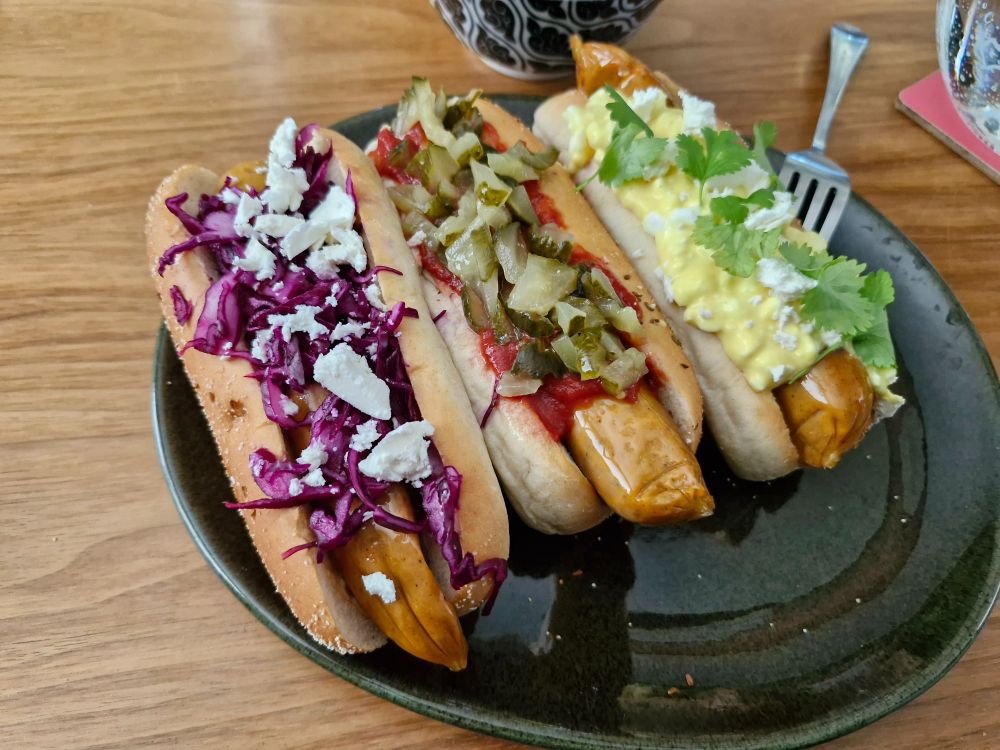 Tre vegokorv med bröd, en med ketchup och inlagd gurka, en med rödkål och vitost och en med mangodressing, keso och koriander som topping.