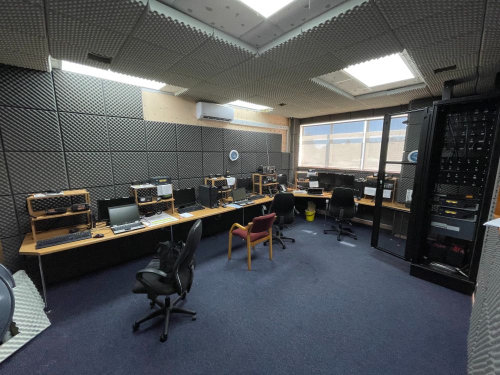 A radio room with a desk along two walls. On top are various ham radios. To the right is a large cabinet which is the patch panels for the antennas and contains battery backup power.