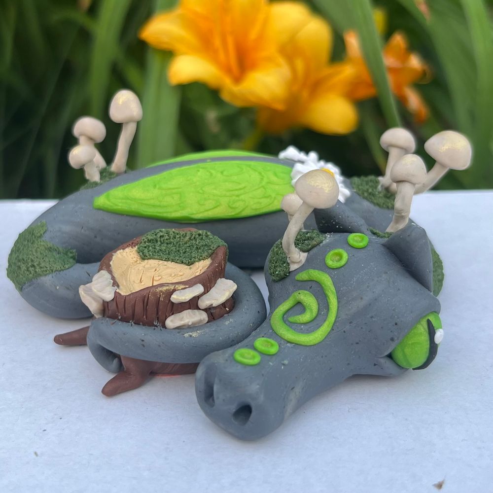 Grey polymer clay dragon with light green eyes, wings, and face swirl/dots, with off-white mushrooms, dark green moss details, and its tail wrapped around a brown/tan tree stump with shelf mushrooms and dark green moss details