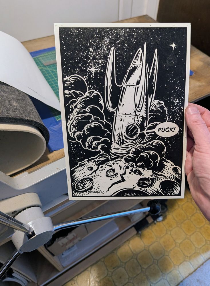 A hand holding up a linoleum print of a crashed space ship. The pilot is saying "FUCK!"