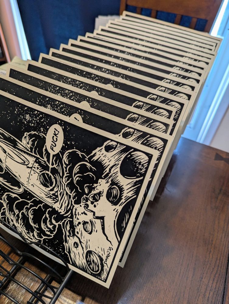 A rack of freshly printed linoleum art prints with the image is a crashed spaceship. The ships pilot is saying "FUCK!"