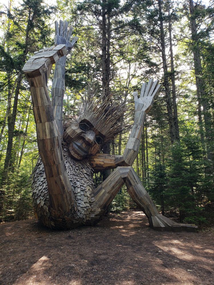 A giant troll made entirely of recycled materials in the forest. The troll is sitting, arms and one leg extended to the sky.