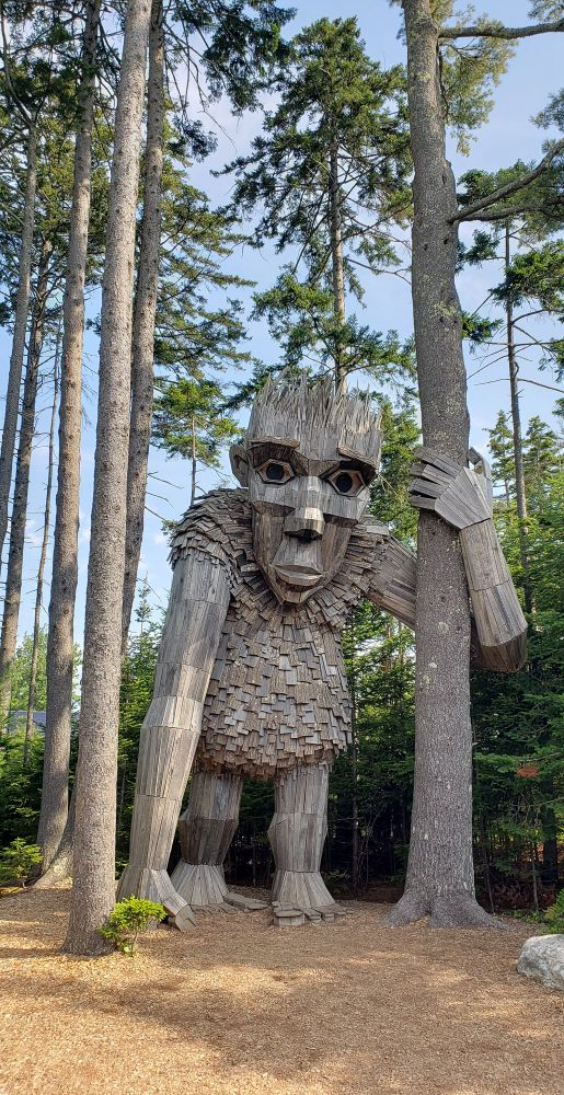 A giant troll made of recycled materials standing with one are wrapped around a tree trunk.