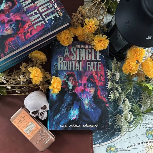 A copy of A SINGLE BRUTAL FATE, a YA fantasy novel with two trans-masc sorcerers on the cover. The book is surrounded by flowers, skulls, a lantern, and various clutter, along with several other stacked copies of the same book.