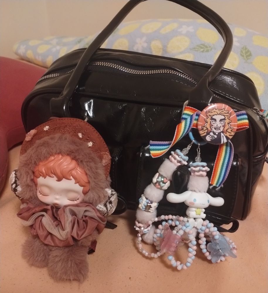 Black handbag with a red skullpanda plush, a Cinnamoroll charm and a rainbow ribbon with a Rengoku Kyoujurou batch