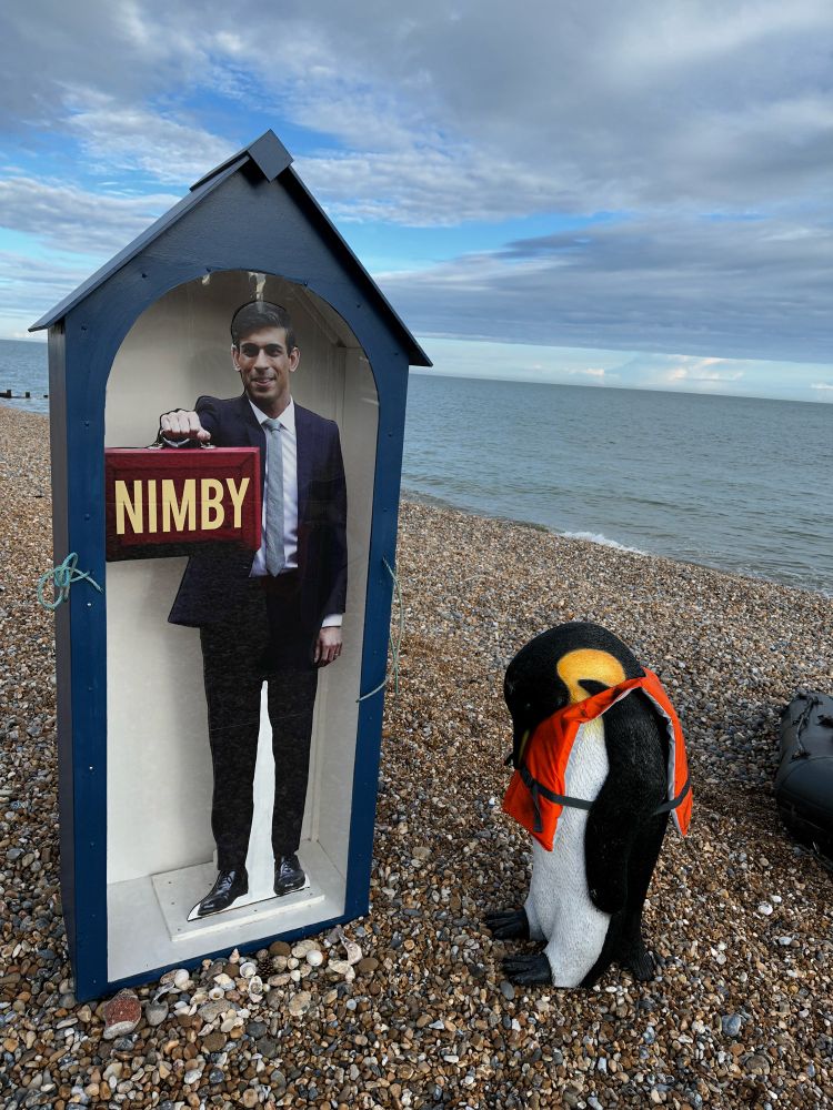 Penguins in life jackets on a dinghy in a life boat next to a cut out in a box saying Not In My Back Yard