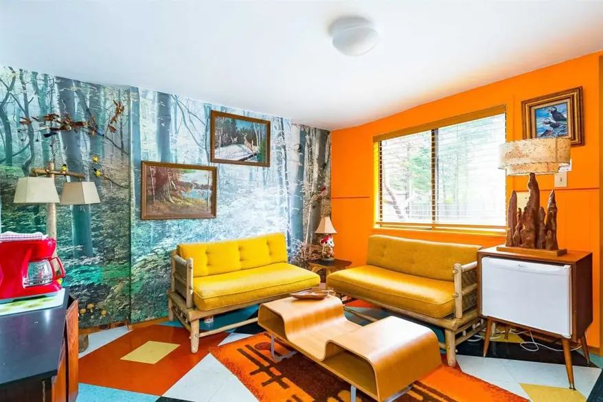 Lounge area with yellow sofas, wallpaper depicting a forest scene, and a bright orange wall on which a framed painting of some puffins hangs