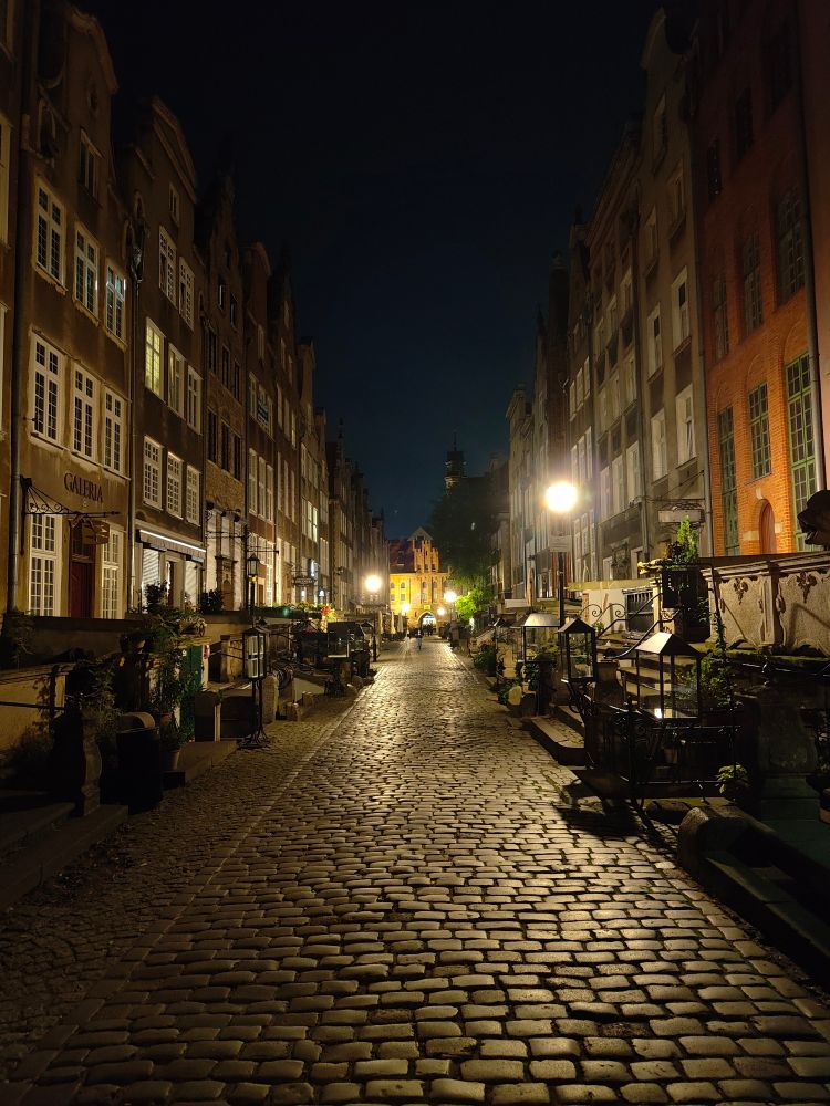 Picture of a street in Gdansk Old Town at night. Warm and neutral lighs bounce off of the cobblestone roads and imposing tight corridor buildings on both sides. With an archway in the far end. Little people can be seen walking in the back.