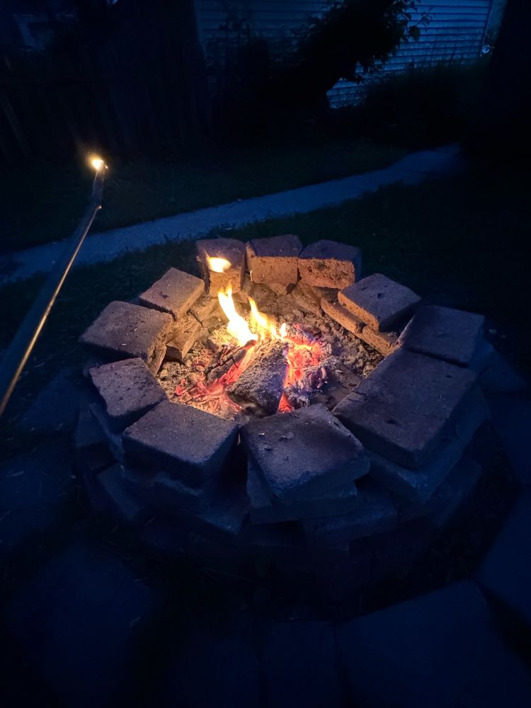 The same fire pit but at night, the fire has died down and is almost out. I’m holding a stick outward, the tip is on fire a small bit.