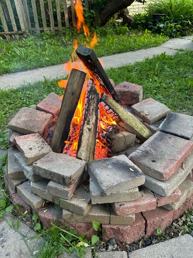 A pic of my back yard with a brick fire pit with logs arranged in a pyramid kind of way, the fire Is consuming the whole thing and it’s beautiful.