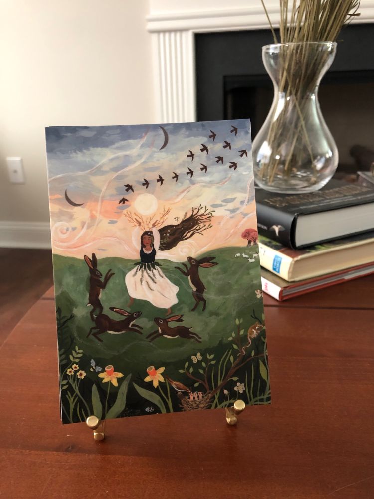 Card sitting on a brass easel showing a woman celebrating spring by dancing with hares. Spring flowers and a bird's nest are in the foreground. The sky in the background shows the sun, two moons, and birds in flight.