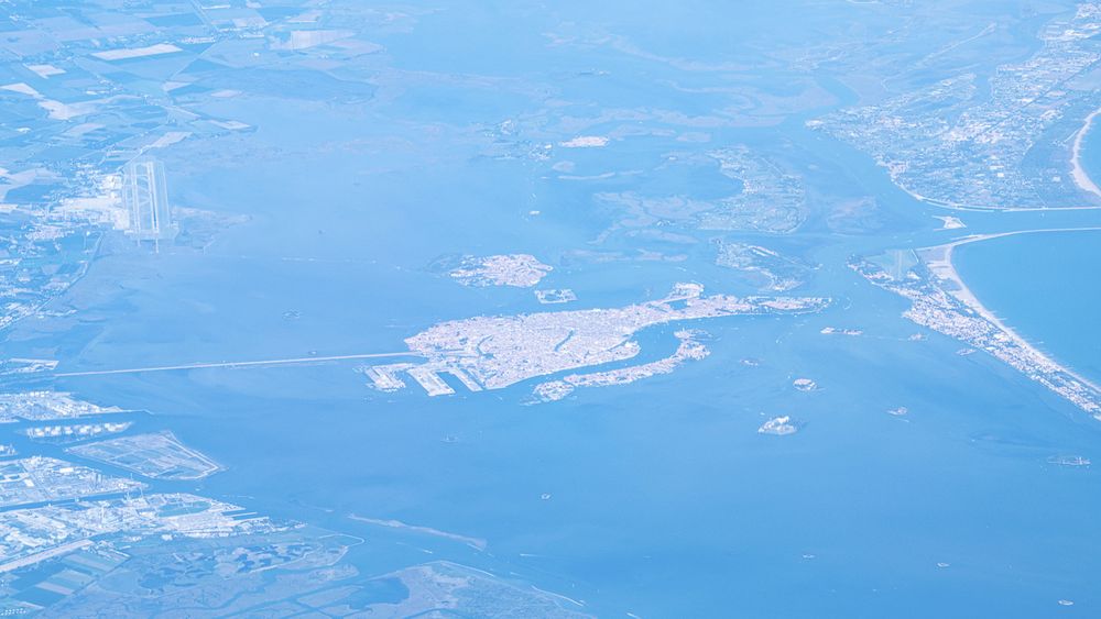 Venice seen from a plane. A large part of the lagoon can be seen, although it is a very similar blue-green to the surrounding land. Venice is a mass of white buildings with red roofs, and some of the larger canals can be seen (as can the causeway connecting the city to the mainland)