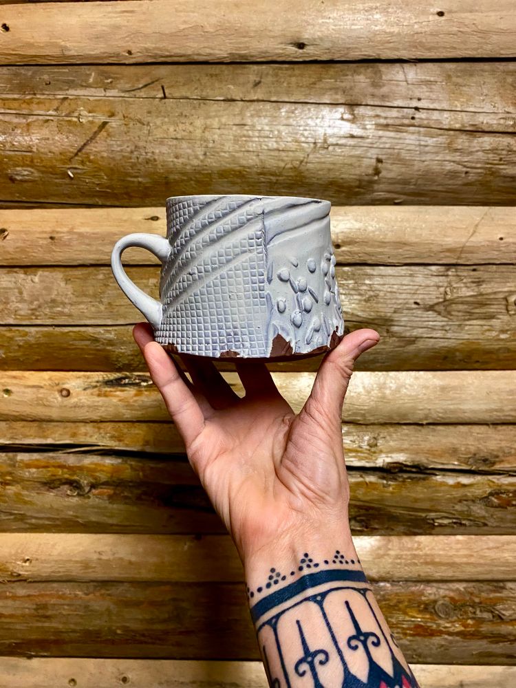 Hand holding an unfired, handbuilt and impression textured mug in front of a log cabin wall