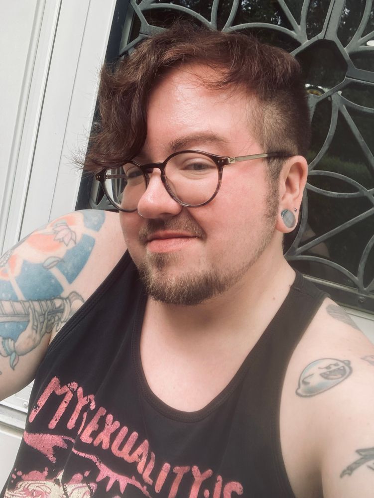A guy with curly auburn hair that is long on top and cascading down the front, but short on the sides; he has several tattoos, round glasses, a mustache and a goatee, and is wearing a black tank top. He has a half smile.