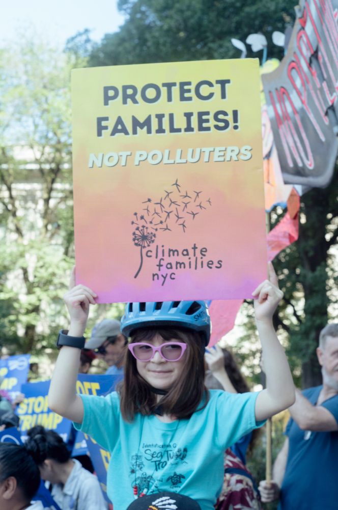 A child in a bike helment and pink glasses holds a sign that says Protect Families, Not Polluters