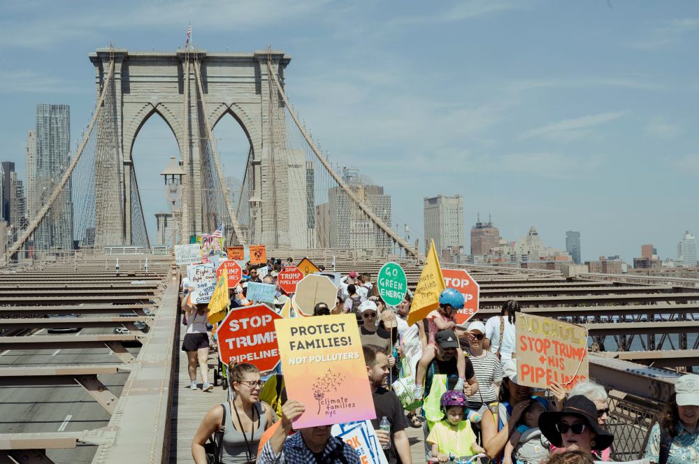 A crowd of kids and adults marching in protest over the Brooklyn bridge, the arches are behind the crowd with the skyline behind, at the front:  A sign that reads "protect families not polluters" 