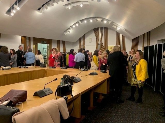 A group of approximately 25 people are standing and chatting in a Scottish Parliament room. The room has a large table with lots of  microphones on it.