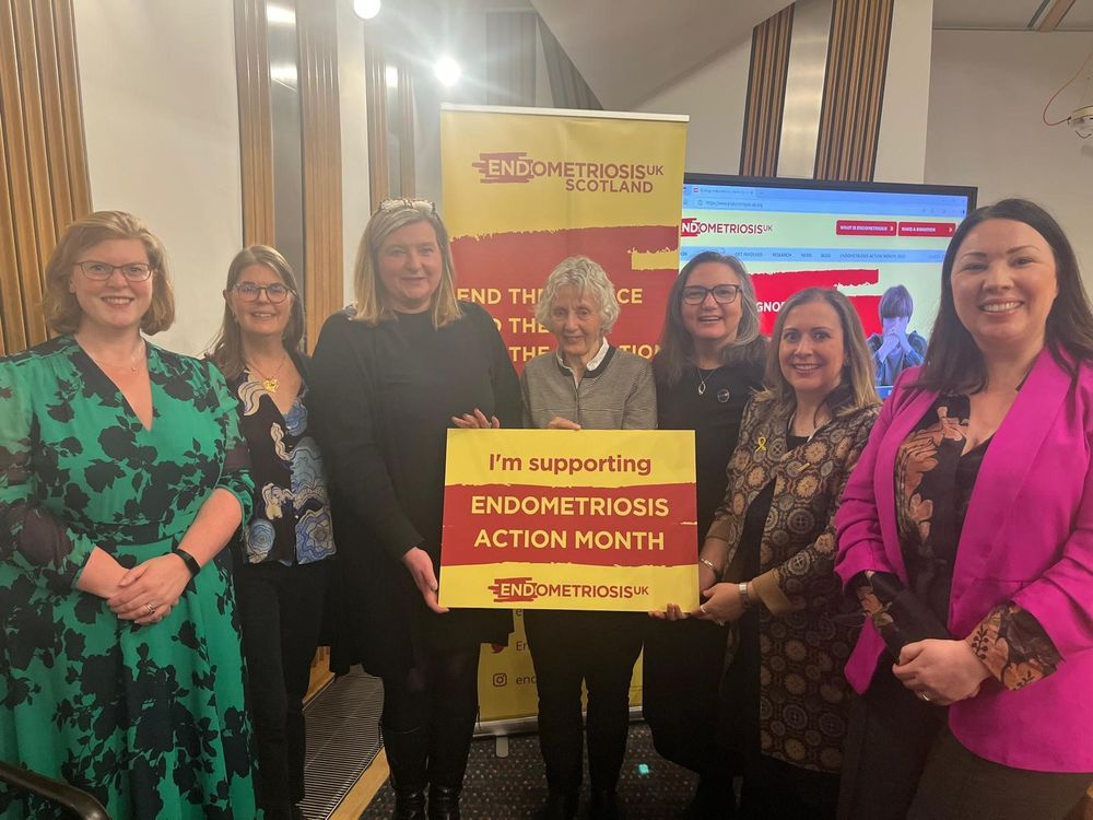 A group of women stand in Scottish Parliament, in front of a banner and holding a board in yellow and red with the words "I'm supporting ENDOMETRIOSIS ACTION MONTH, Endometriosis UK". They are smiling at the camera.