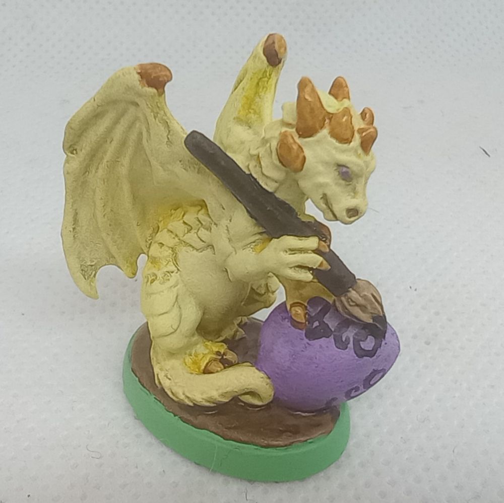 Figurine of a young yellow dragon painting silly faces on a pastel purple egg