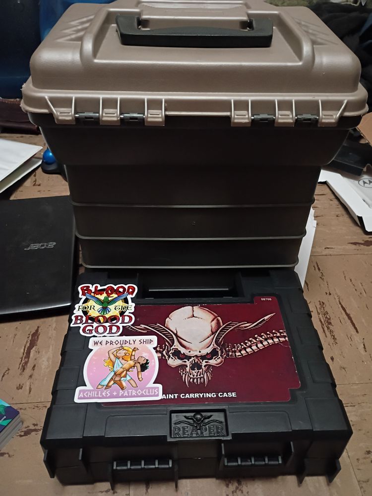 Picture of the back of a tackle box and paint case in desperate need of stickers. The tackle box is woefully barren. The paint case has the Reaper Miniatures logo, and 2 stickers by Red of Overly Sarcastic Productions, a "We proudly ship Achilles and Patroclus" and Huitzilopochtli "Blood for the blood god" hummingbird.
