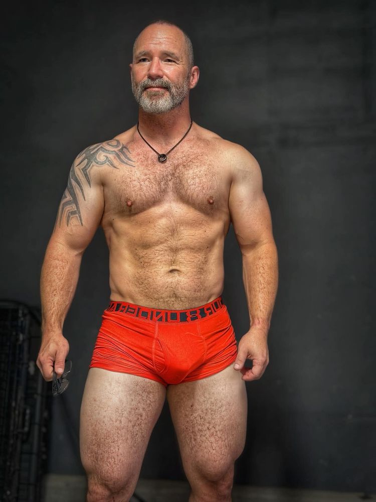 The same beefy muscular man stands wearing only red Under Armour boxer briefs