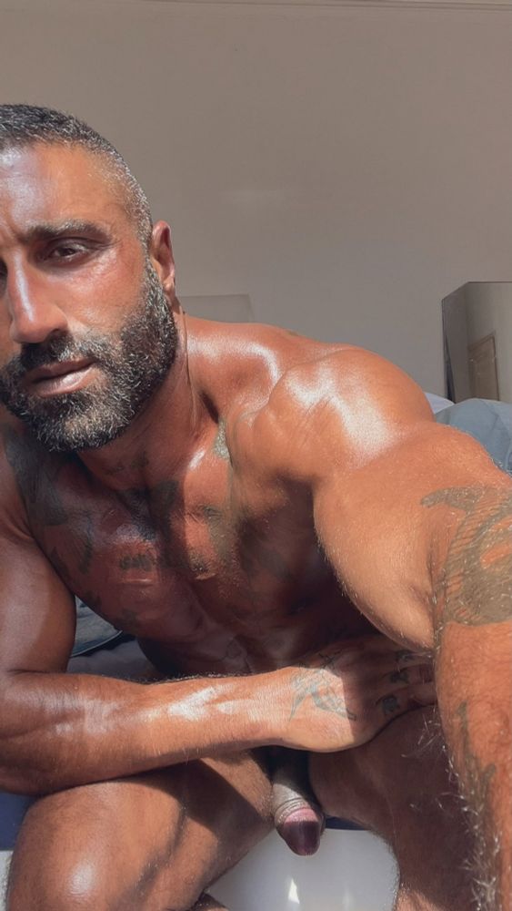 A handsome, muscular Middle Eastern man with a tan and tattoos sits naked, leaning towards the camera