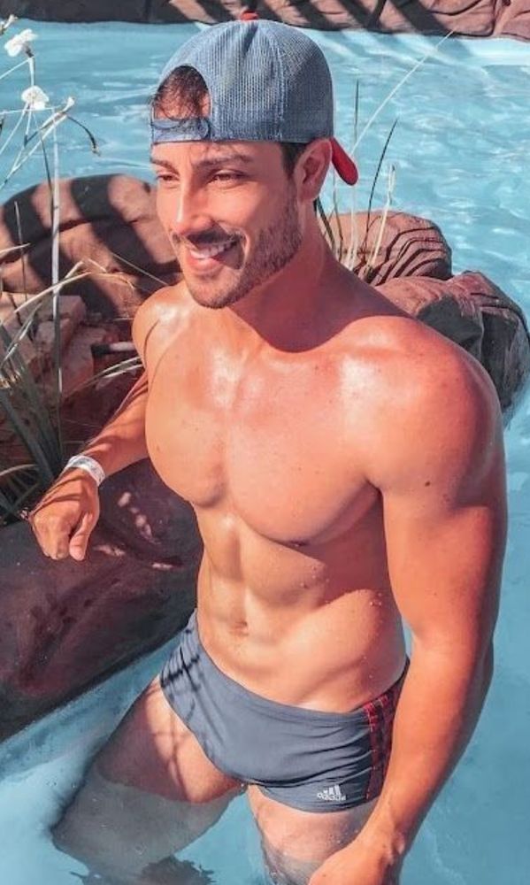 A handsome, muscular young man stands in a swimming pool, wearing gray trunks, a backwards ballcap and a smile