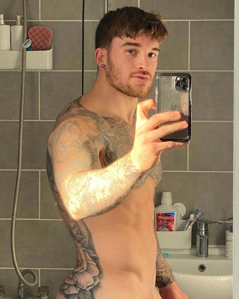 A handsome muscular tattooed man takes a side-on naked selfie in his bathroom