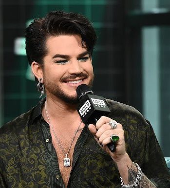Screenshot from an interview with Adam Lambert in 2019, holding the show microphone and smiling, in a dark green paisley print shirt.