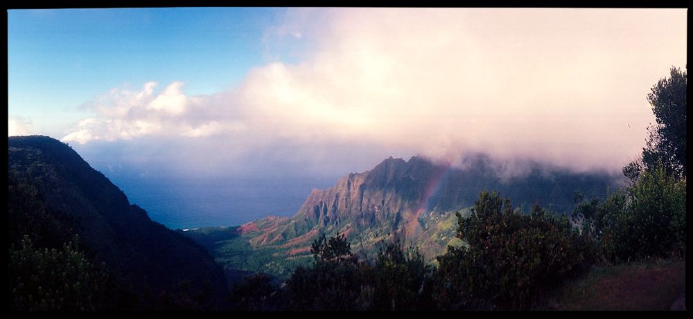 Panoramic shot of tropical cliffs in the distance, ocean beyond them, soft white clouds moving in, and a bit of a rainbow against the cliffs.