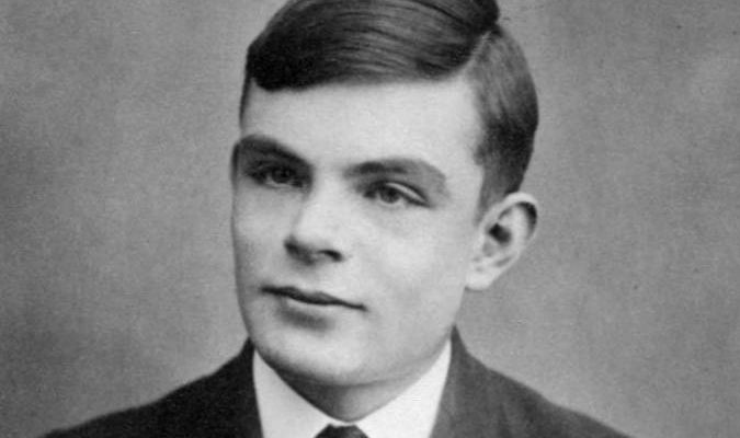 A black and white photograph of a young Alan Turing, looking thoughtfully off to the left, wearing a jacket, shirt and tie, and with a side parting.