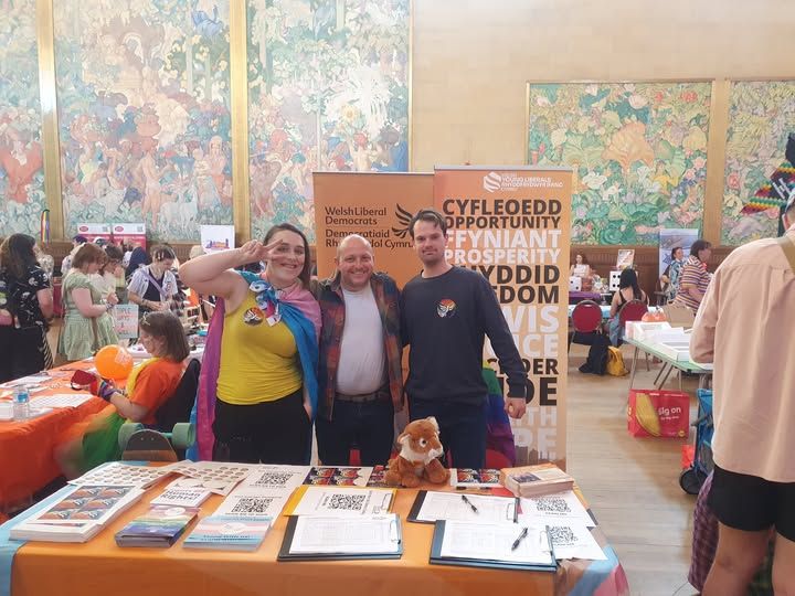 Three people standing smiling behind an amber-clad table in an airy hall with tapestries in the background. Behind them are Liberal Democrat banners in Welsh and English; the table is scattered with stickers, QR codes, leaflets and petitions to sign, as well as a plushy tiger. The woman on the left is wearing a yellow top with a trans flag on top worn as a cape; she and the man on the right are wearing rainbow stickers with the Lib Dem bird-of-liberty logo on.
