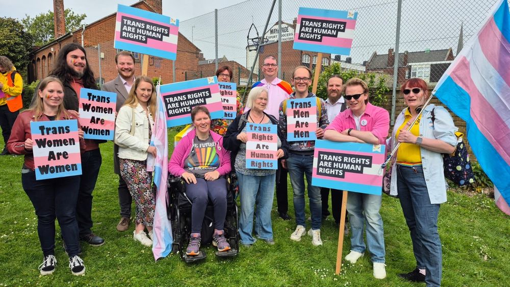 A picture of a diverse group of smiling Liberal Democrats waving placard in the colours of the trans flag, saying 'Trans Women Are Women', 'Trans Men Are Men', and 'Trans Rights Are Human Rights'.  Most of the placards have a rainbow Lib Dem bird-of-liberty in the corner, and the woman on the right is carrying a very large trans flag on a pole.  In the centre of the group are an older woman and a smiling woman in a wheelchair.