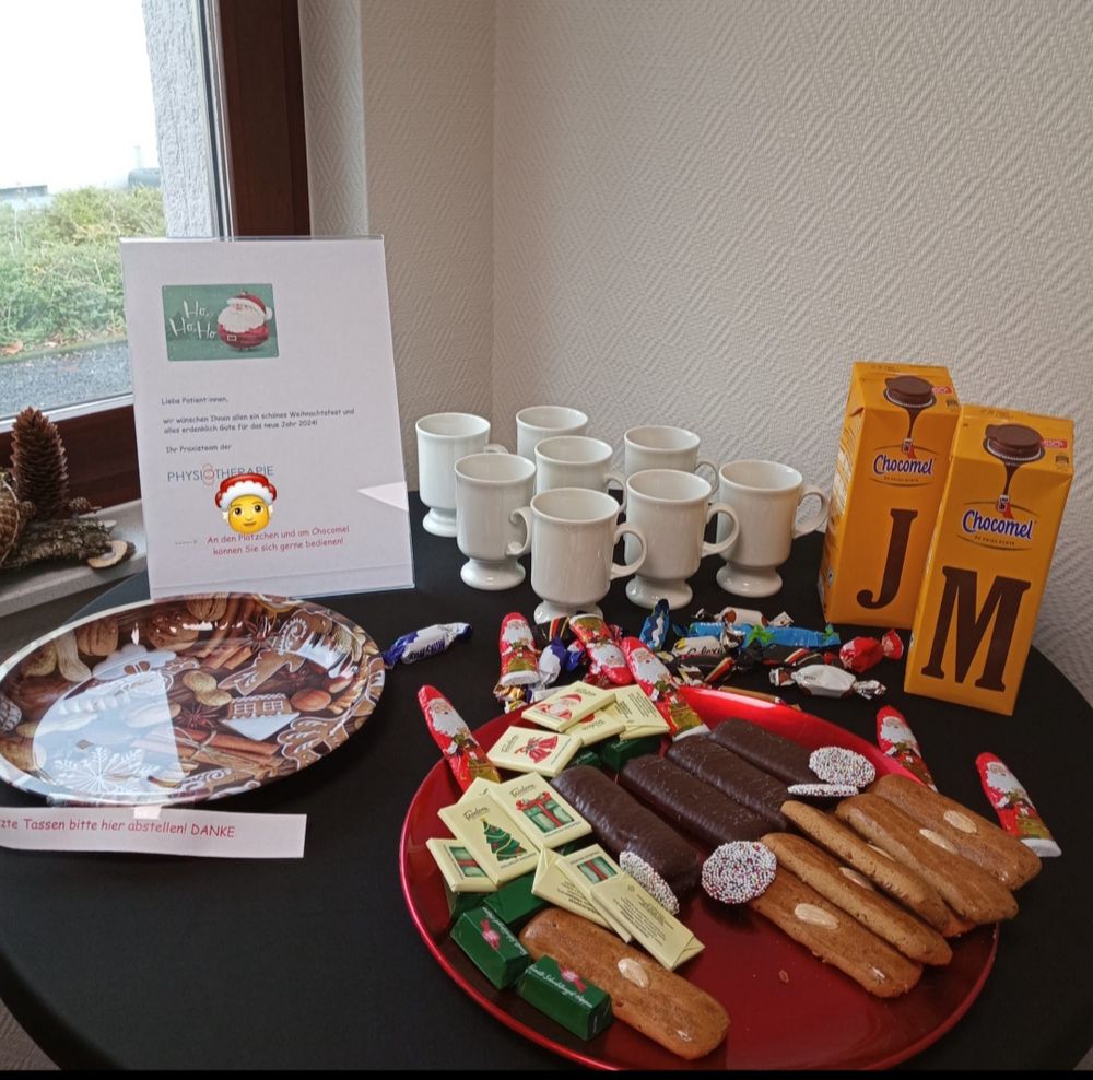 Ein Stehtisch mit einem Weihnachtsteller ,auf dem verschiedene Weihnachts Naschereien sind . Im Hintergrund größere Tassen und Packungen mit Choccomel Kakao. 
Mit anderen f dem Tisch ein Schild ,auf dem allen Patienten der Physiotherapie Praxis Frohe Weihnachten gewünscht wird ,und die Bitte,sich an den Leckereien zu bedienen .
