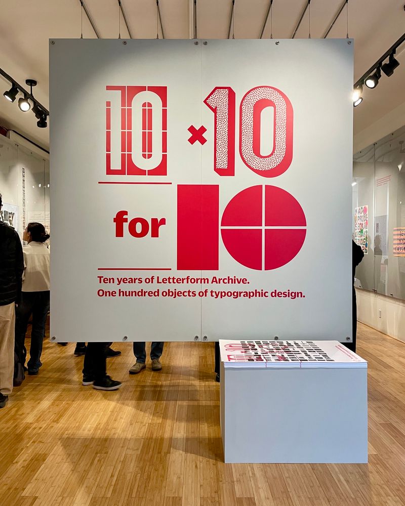 Photo of a wood-floored gallery with a large floating gray wall filling most of the frame. In the wall are large red vinyl letters: “10 × 10 for 10” in different styles, and “Ten years of Letterform Archive. One hundred objects of typographic design.” in smaller text. A stack of posters on a pedestal is in the foreground, and people looking at art in the gallery in the background. 