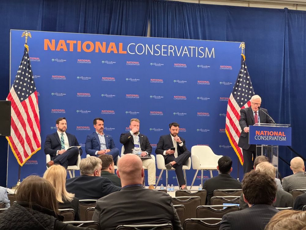 Panelists sit in seats on a NatCon panel as a person introduces them from the podium/lectern.