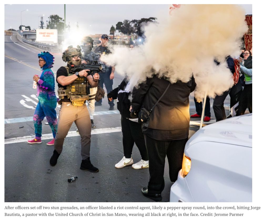 A pastor stands in the road, his head engulfed in yellow smoke fired by a DHS officer who stands just a few feet away.