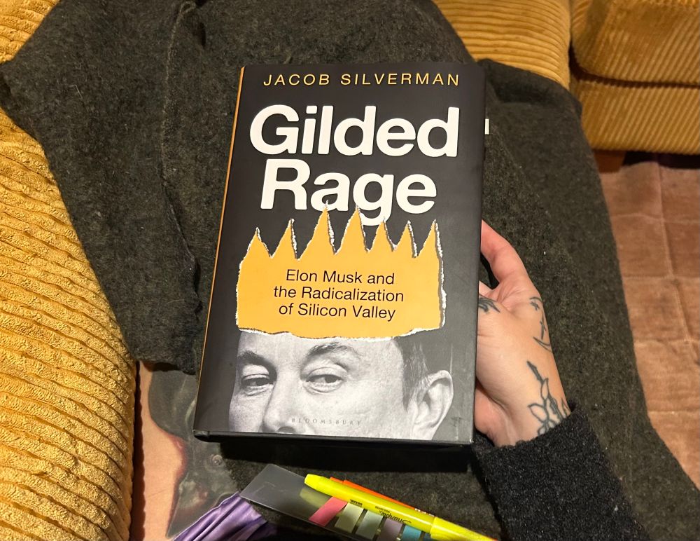 A book on Alyssa’s lap. Gilded Rage: Elon Musk and the Radicalization of Silicon Valley by Jacob Silverman. Half of Musk’s face is on the cover. There’s a highlighter and Post It tabs on her map, too. 