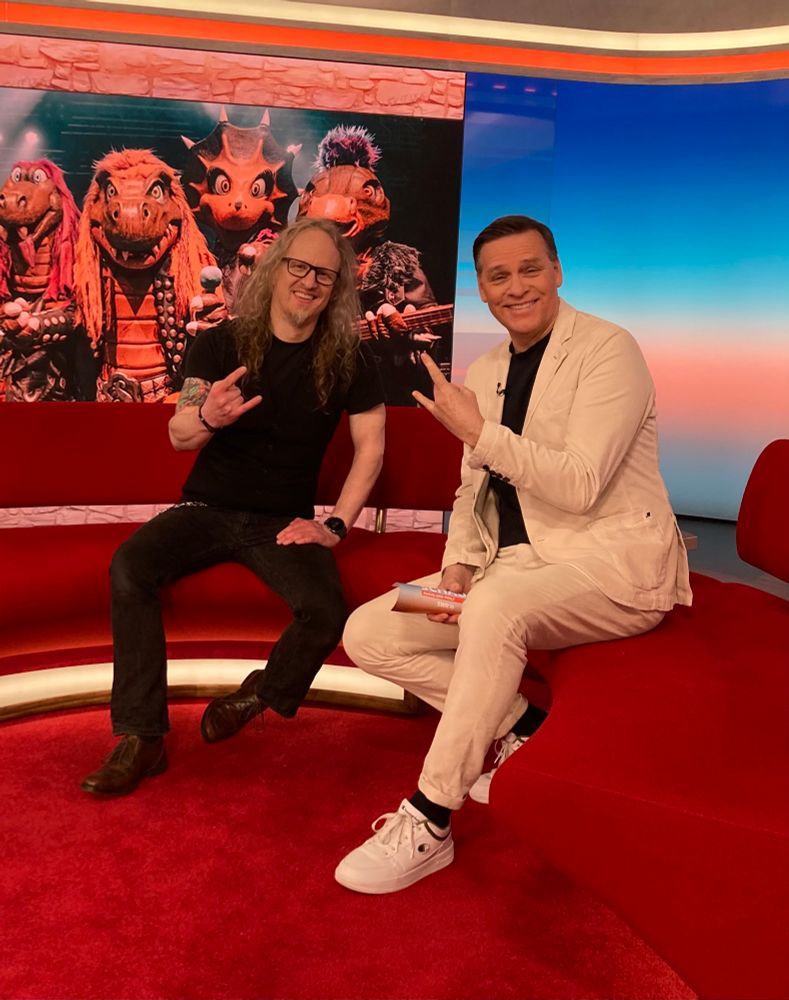 Musiker & Journalist Christof Leim mit Moderator Holger Wienpahl im Studio der SWR Landesschau Rheinland-Pfalz. Im Hintergrund ein großes Bild von Heavysaurus. 