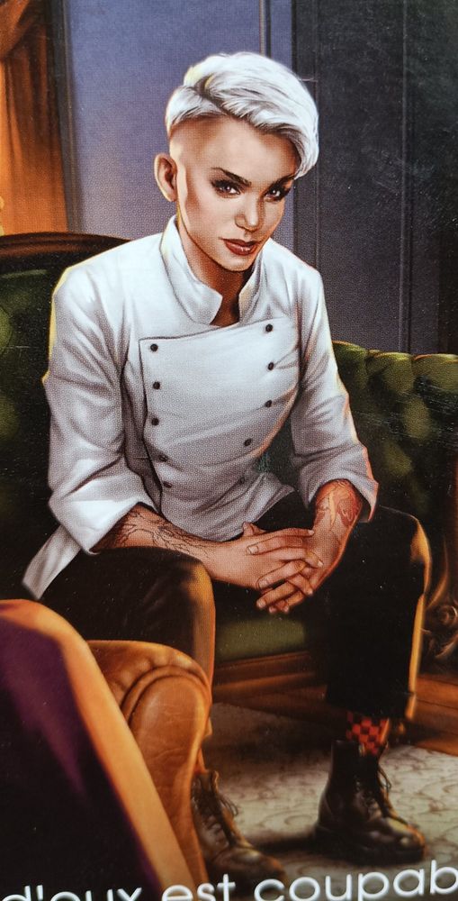 Un personnage de la nouvelle édition du Cluedo, la Cheffe Leblanc, assise dans un fauteuil. Elle est en pantalon, veste de cheffe de cuisine, elle a les cheveux courts, teintés en blanc avec un side-cut