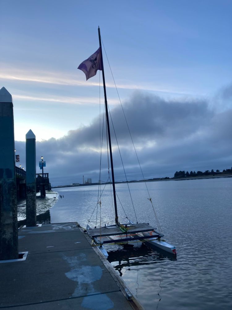 A photo of a pontoon raft tied to a dock on Humboldt Bay in Eureka, CA. There is a TIE DYED LOL :) pirate flag flying from a staff that looks to be 10 to 20 ft. (~ 3 - 7 meters); which is planted in the middle of the raft. I took this pic in 2024 or earlier.