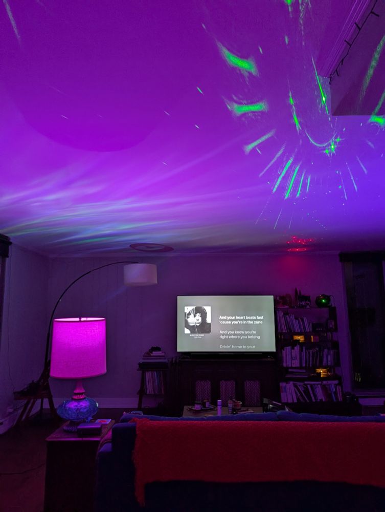 Living room bathed in purple and blue light, with lasers and aurora projections on the ceiling and Lady Gaga's new album playing on the TV