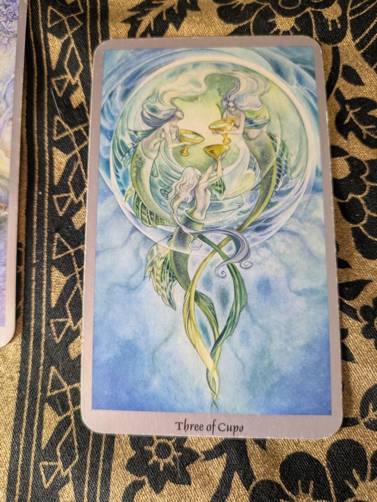 A tarot card rests on a gold and black cloth. It is the Three of Cups. On its face are three mermaids swimming in a circle so quickly that they've created a large air bubble just for themselves. At the center of the circle they joyfully offer their cups to one another. 