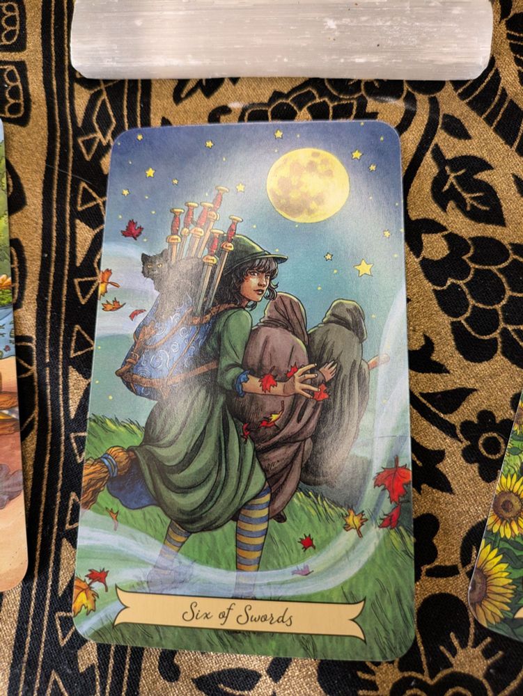 On a gold and black table cloth rests a tarot card. The card is labeled the Six of Swords in scrolled letters on a cream pennant at the bottom of the picture. A young, dark haired witch rides a broom behind two robed friends. In her blue-scrolled backpack are six swords and a black cat, which might be too much to handle by herself on what is obviously a very windy day; leaves are blowing about and the wind itself is depicted as a translucent jet of air in the foreground. The witch and her friends are about to fly over a body of water, and a distant shore is just visible in the background. Above them, a huge full moon rides high in a sky full of stars. 