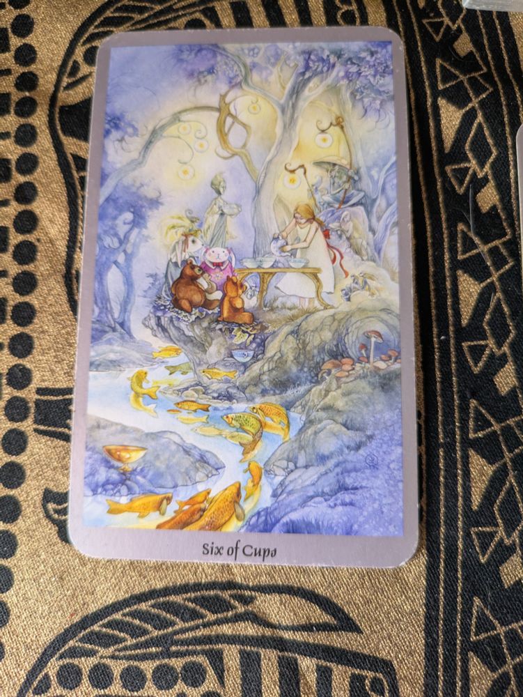 A tarot card rests on a gold and black floral cloth. It is the Six of Cups. On its face a young woman pouring tea into rather large cups on a small table. She is in the woods, surrounded by fairies and sprites--some of which hold cups--and before the table are stuffed animals which could arguably be said to look alive. Through the forest flows a stream teeming with fish. 