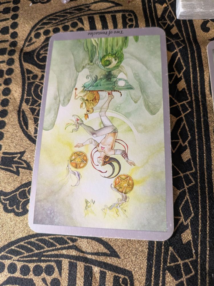A closeup of the first tarot card, this image shows a whimsical man dressed like a mime or an acrobat balancing on one foot atop a small pedestal while juggling two golden pentacles above his head. The card is upside down. 