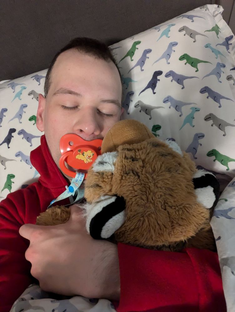 A photo of me in bed from my chest up. I'm lying on my back with my head on the pillow and my eyes closed. There's a big red pacifier with a lion printed on it in my mouth. My arms are wrapped around my big tiger plushie, but you can only see the back of his head here because the rest of him is under the blankie. His muzzle is right up against my cheek. I love snuggling like this!