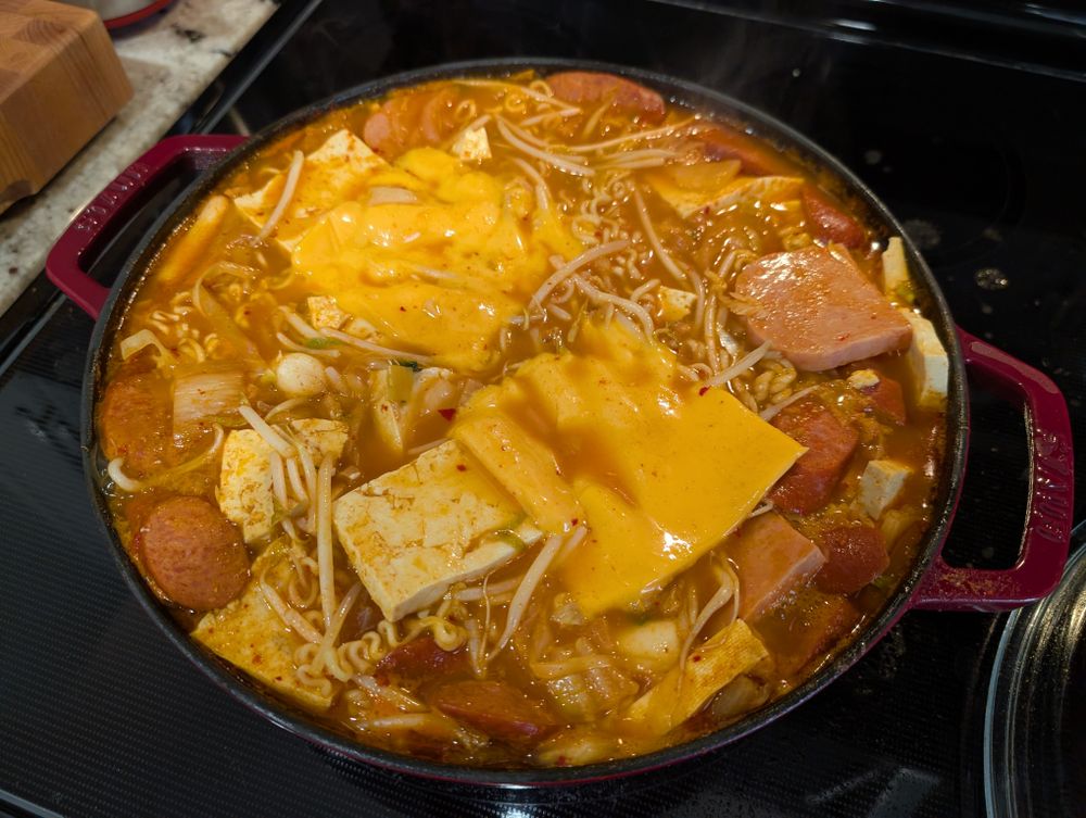 My budae jjigae on the stove! It's Korean "Army Base Stew" and it has everything in it. Mine has cabbage, onions, green onions, kimchi, spam slices, hot dog slices, tofu, ramen noodles, bean sprouts, and processed cheese. The photo shows my stew with pretty much all of those ingredients in an orange broth, which is in a wide and shallow enamel cast iron pot that is full right to the brim (it got a little spilly).