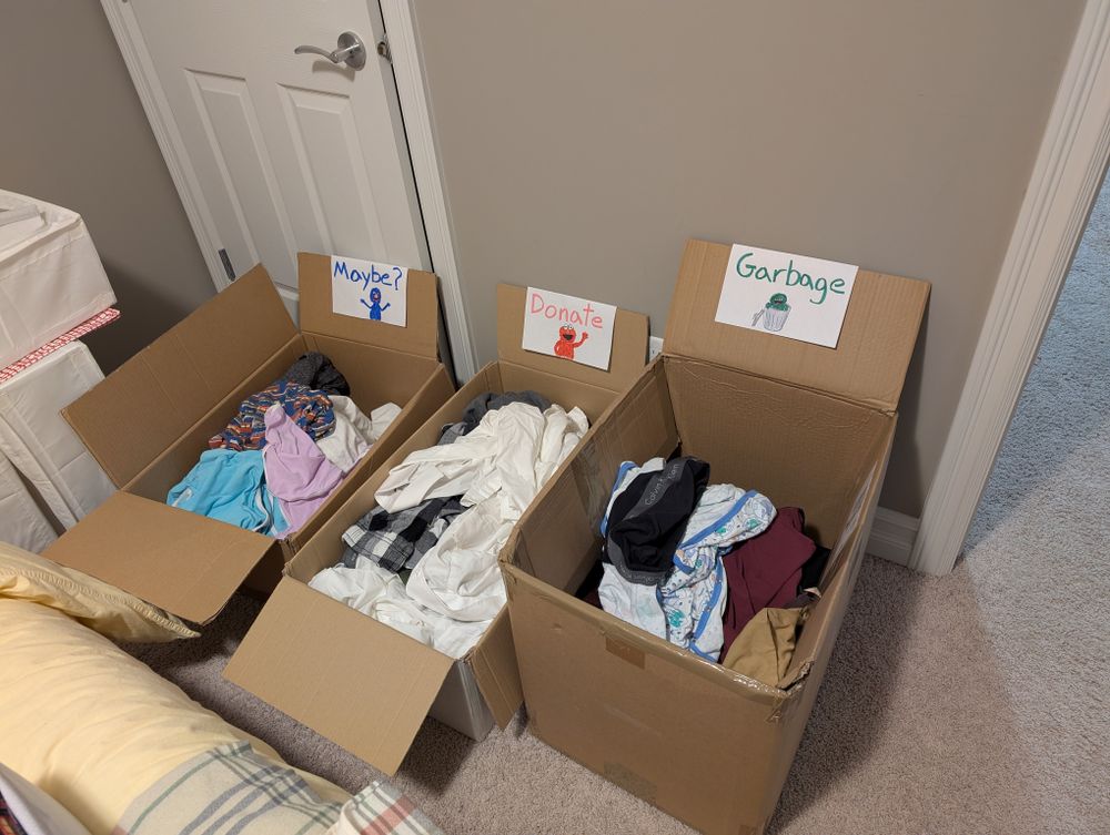 A photo of three large boxes that may have previously contained diapers sitting on the floor side by side. They each have one of the top flaps propped against the wall with a hand drawn label on it.