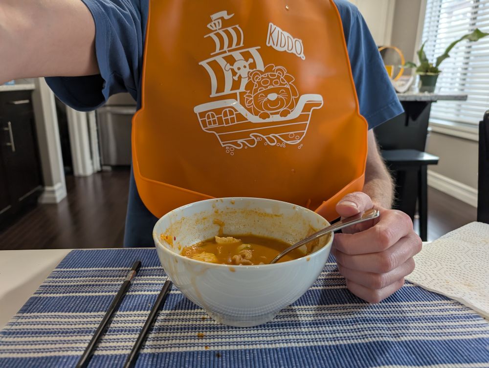 Dinner time! This is a photo of me from across the table, showing me wearing an orange silicone bib as I eat a bowl of stew.